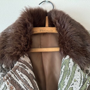 K&ouml;nnte beinhalten: Ein brauner Pelzkragen an einer Jacke mit gr&uuml;nem und wei&szlig;em Muster. Die Jacke h&auml;ngt an einem Holzb&uuml;gel mit dem aufgedruckten Text "For Presidio".