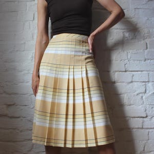 May include: A high-waisted, knee-length pleated skirt featuring a horizontal plaid pattern in shades of pale yellow, white, and thin black lines. The skirt has a structured waistband and a series of vertical pleats that extend from the waist down.