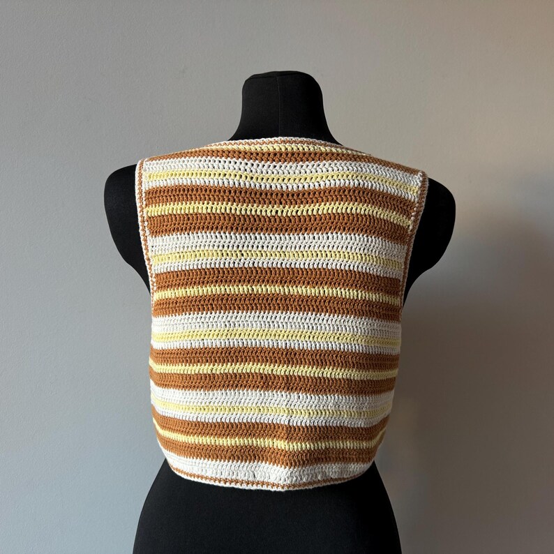 Puede incluir: Un top de crochet a rayas marrones y blancas con cuello redondo. El top es sin mangas y corto.