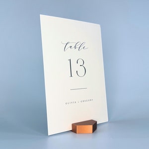 May include: White card table number holder with the text "table 13" and "Olivia + Gregory" printed on it. The card is sitting on a copper-colored stand.