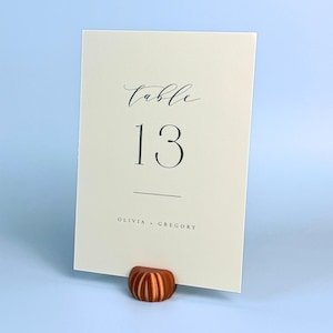 May include: A white card with black calligraphy that reads "table 13" and "Olivia + Gregory". The card is held by a brown wooden table number holder.