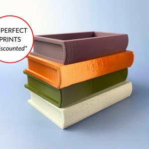 May include: A stack of four 3D printed book-shaped boxes. The top box is purple, the second is orange, the third is green, and the bottom box is white with a speckled texture. The text "IMPERFECT PRINTS *discounted*" is visible in a red circle.