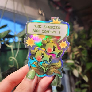 Puede incluir: Una pegatina holográfica con dos plantas de dibujos animados verdes con el texto "The Zombies Are Coming!" en un bocadillo. La pegatina tiene un acabado iridiscente arcoíris.