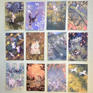 Op de afbeelding: Een verzameling van twaalf geïllustreerde kunstprints. Elke print toont grillige scènes met dieren, waaronder konijnen, katten en eenhoorns, tegen kleurrijke achtergronden met sterren, bomen en water. De prints hebben een dromerig fantasiethema.