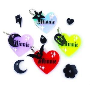 May include: Assortment of colorful heart-shaped keychains with the word "Minnie" on each. The keychains feature different charms, including a star, a crescent moon, a bat, and a skull. The charms are black and the hearts are iridescent, purple, yellow, and red.