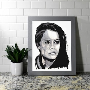 May include: A black and white portrait of a woman in a gray frame. The artwork features a detailed depiction of a woman's face, with expressive eyes and dark hair. The portrait is set against a white background.