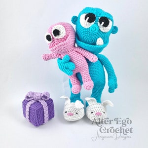 May include: A blue crocheted monster holding a pink crocheted monster. The blue monster is wearing white bunny slippers. A purple crocheted gift box is in the foreground. The text "Alter Ego Crochet Amigurumi Designer" is in the bottom right corner of the image.