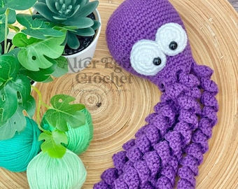 May include: A handmade purple crochet octopus with white eyes and black pupils. The octopus is lying on a wooden plate, with green yarn and a potted succulent plant in the background. The octopus is a whimsical, handcrafted item.