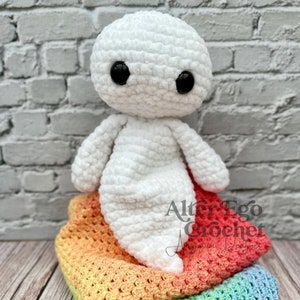 Könnte beinhalten: Ein weißes gehäkeltes Geister-Amigurumi-Spielzeug mit schwarzen Augen, das auf einer regenbogenfarbenen gehäkelten Decke sitzt. Die Decke ist in drei Teile gefaltet, wobei die oberste Schicht pfirsichfarben, die mittlere Schicht hellgrün und die unterste Schicht blau ist. Die Decke liegt auf einer Holzoberfläche.