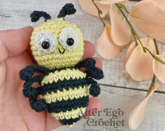 May include: A crocheted amigurumi bee with yellow and black stripes, white eyes, and black pupils. The bee is being held in a hand with a light pink flower behind it.