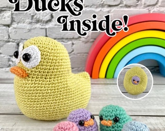 Puede incluir: Un conjunto de cinco peluches de pato de crochet. El pato más grande es amarillo y tiene un arcoíris detrás. Los patos más pequeños son rosa, morado, verde y azul.