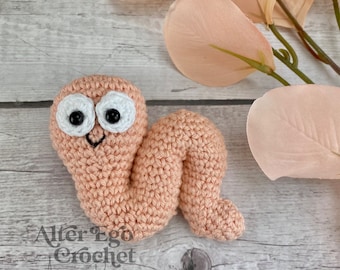 Op de afbeelding: Een gehaakte roze worm met witte ogen en een zwarte glimlachende mond. De worm ligt op een lichte houten achtergrond met roze bloemstengels.