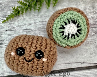 May include: Two crocheted kiwi fruit, one whole and one sliced, with a smiling face on the whole kiwi. The sliced kiwi features green, brown, and white yarn. The whole kiwi is brown with black eyes and pink cheeks. The background is a wooden surface.