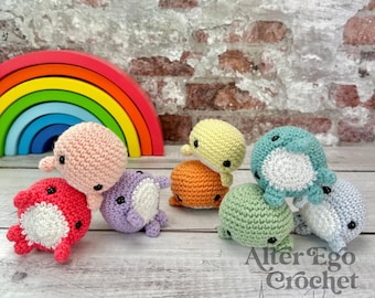 May include: A collection of colorful, crocheted whale amigurumi toys in various colors, including pink, red, purple, yellow, orange, green, and blue. A wooden rainbow and the text "Alter Ego Crochet Amigurumi Designer" are in the background.