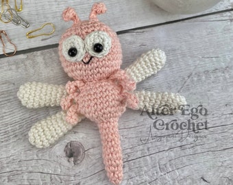 May include: A pink and white crocheted dragonfly amigurumi toy with black eyes. The toy is lying on a light wood surface. The text "Alter Ego Crochet Amigurumi Designer" is visible in the lower right corner of the image.