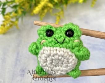May include: A small green crocheted frog with black eyes and a white belly, sitting on a wooden stick. The frog is a handmade amigurumi toy.