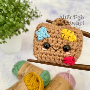 Op de afbeelding: Een bruin gehaakt amigurumi-speelgoed met een glimlachend gezicht, blauwe en gele plekken en een rode lip. Het speelgoed wordt vastgehouden door houten eetstokjes. Er zijn vier bollen garen op de achtergrond: geel, groen, roze en wit.