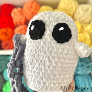 Puede incluir: Juguete amigurumi fantasma de crochet blanco con ojos negros y una cuerda de crochet gris. El juguete está sentado en un estante con ovillos de lana de colores.