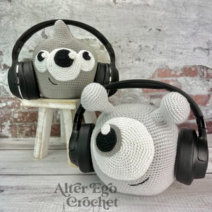 Può includere: Due figure amigurumi all'uncinetto grigie e bianche con grandi occhi e cuffie nere. Le figure sono sedute su una superficie di legno. Il testo "Alter Ego Crochet" è scritto sotto le figure.