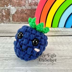 Peut inclure: Un jouet amigurumi en crochet en forme de myrtille avec un visage souriant, des yeux noirs et des joues roses. Le jouet est posé sur une surface en bois avec un jouet arc-en-ciel en arrière-plan. Le texte "Cuter Ego Crochet Amigurumi Designer" est visible dans le coin inférieur droit de l'image.