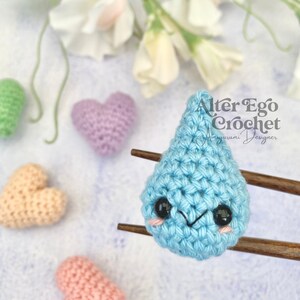Puede incluir: Una gota de lluvia de crochet azul con una cara linda, descansando sobre palillos de madera. La gota de lluvia está rodeada de corazones de crochet en varios colores.