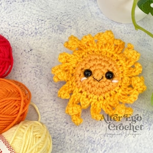 Puede incluir: Un sol amarillo tejido a crochet con una cara sonriente y ojos negros. El sol está sobre una superficie blanca texturizada con ovillos de lana en el fondo. El texto "Alter Ego Crochet Amigurumi Designer" es visible en la esquina inferior derecha de la imagen.