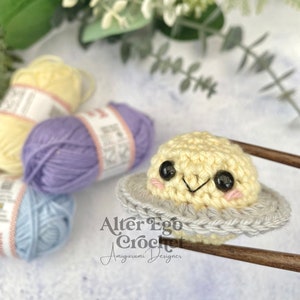 May include: A crocheted amigurumi toy of a yellow planet with a gray ring around it. The planet has a smiling face with black eyes and pink cheeks. The toy is being held by chopsticks. The text "Alter Ego Crochet Amigurumi Designer" is visible in the image.