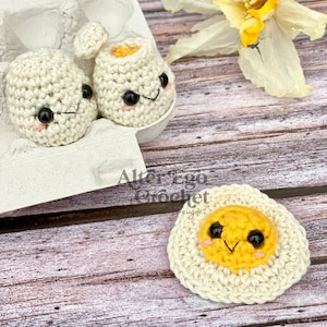 May include: Three crocheted eggs, two in an egg carton and one on a wooden surface. The eggs have cute faces and the one on the surface is cracked open to reveal a yellow yolk.