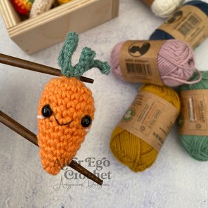 May include: A crocheted carrot amigurumi with a smiling face and green leaves. The carrot is sitting on a pair of wooden chopsticks. There are several balls of yarn in the background, including white, pink, blue, green, and yellow.
