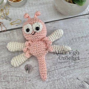 May include: A pink and white crocheted dragonfly amigurumi toy with black eyes. The toy is lying on a light wood surface. The text "Alter Ego Crochet Amigurumi Designer" is visible in the lower right corner of the image.
