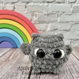 May include: A gray crocheted owl amigurumi toy with large black eyes and white accents. The owl is sitting on a wooden surface in front of a rainbow-colored wooden arch.