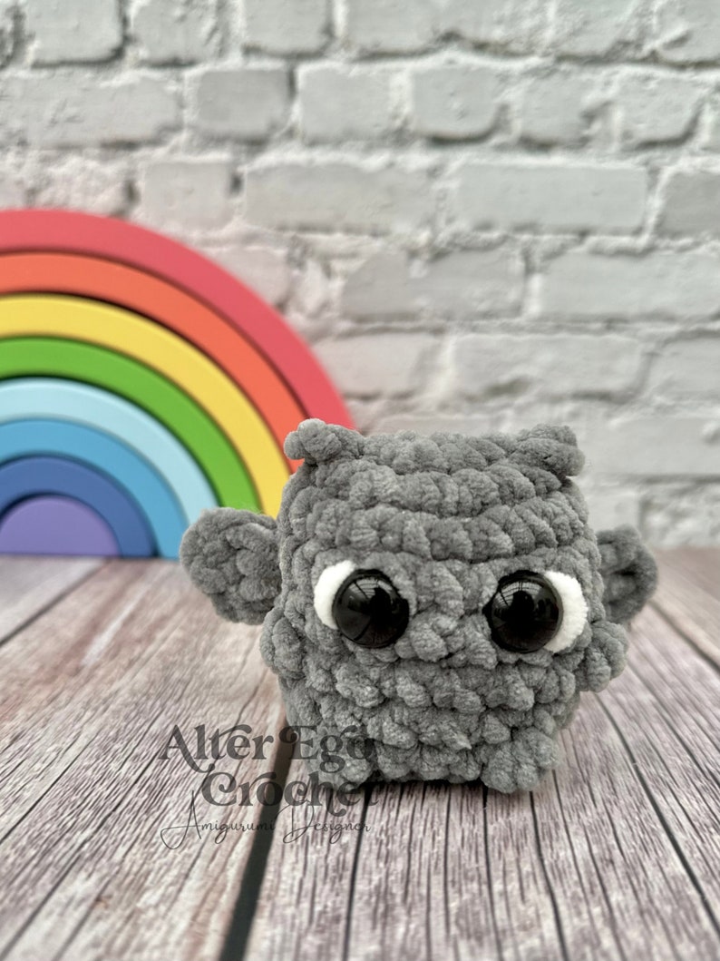 May include: A gray crocheted owl amigurumi toy with large black eyes and white accents. The owl is sitting on a wooden surface in front of a rainbow-colored wooden arch.