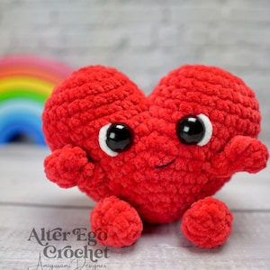 Könnte beinhalten: Ein rotes, gehäkeltes Herz-Amigurumi-Spielzeug mit einem lächelnden Gesicht und großen schwarzen Augen. Das Spielzeug hat kleine Arme und Beine.