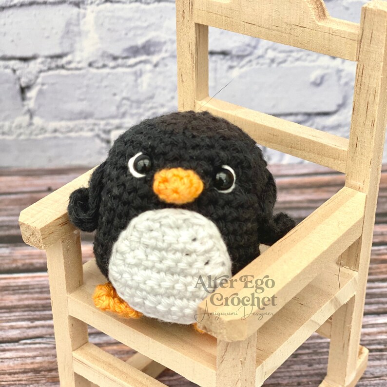 May include: A small crocheted penguin with black and white fur, orange beak, and black eyes sits on a wooden rocking chair. The penguin is wearing a white belly and has a small orange foot visible. The chair is made of light brown wood and has a white background.