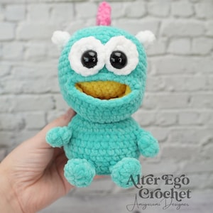 Puede incluir: Un monstruo amigurumi turquesa hecho a mano con grandes ojos con borde blanco, pupilas negras y boca amarilla. Tiene pequeños cuernos blancos, un cuerno rosa en la parte superior y pequeños brazos y pies. El texto "Alter Ego Crochet Amigurumi Designer" es visible.
