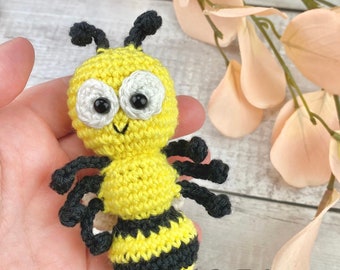 May include: A crocheted bee amigurumi toy with black and yellow stripes, white eyes, and black antennae. The bee is being held in a hand.