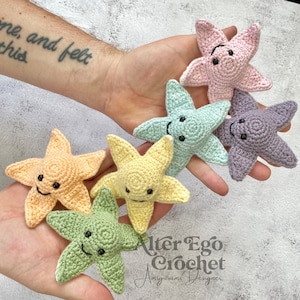 May include: Five crocheted starfish in different pastel colors, each with a smiling face. The starfish are arranged in a semi-circle in the palm of a hand. The text "Alter Ego Crochet Amigurumi Designer" is visible in the bottom right corner of the image.