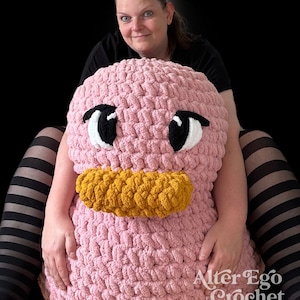 May include: A large pink crocheted plush toy shaped like a duck with black eyes, a white mouth, and a yellow beak. The toy is sitting on a person's lap.