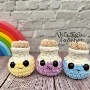 May include: Three crocheted potion bottles in yellow, purple, and blue with white lids and black eyes. The bottles are sitting on a wooden surface. The text "Alter Ego Crochet" is visible in the background.