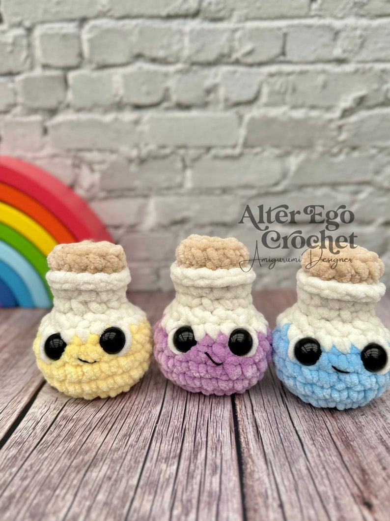 May include: Three crocheted potion bottles in yellow, purple, and blue with white lids and black eyes. The bottles are sitting on a wooden surface. The text "Alter Ego Crochet" is visible in the background.