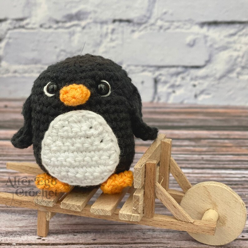 May include: A small black and white crocheted penguin with orange feet and a black beak sits in a wooden toy wheelbarrow.
