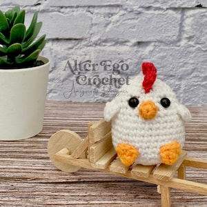 Puede incluir: Pollo amigurumi de crochet blanco con patas naranjas y una cresta roja. El pollo está sentado en un carro de juguete de madera.