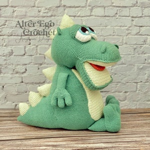 Puede incluir: Juguete de dinosaurio de crochet verde con vientre verde claro y lengua roja. El dinosaurio tiene una expresión triste con ojos grandes y una boca ancha. El juguete está sentado sobre una superficie de madera.
