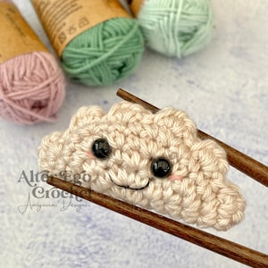 Può includere: Un amigurumi a forma di nuvola all'uncinetto con un viso sorridente e occhi neri. La nuvola è beige ed è tenuta da due bacchette di legno. Lo sfondo è una superficie blu chiaro.