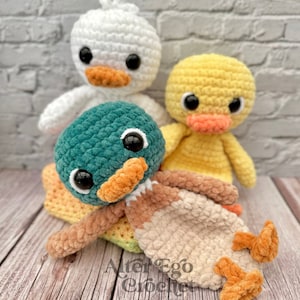 Könnte beinhalten: Drei gehäkelte Enten-Plüschtiere, eine weiße, eine gelbe und eine grüne mit braunen und beigefarbenen Akzenten. Die Enten sitzen auf einer regenbogenfarbenen gehäkelten Decke.
