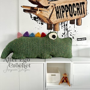 May include: Green crocheted alligator plushie with colorful spikes on its back. The plushie is lying on a white shelf with a wooden monkey toy and a white box in the background. The text "Alter Ego Crochet Amigurumi Designer" is visible in the bottom left corner.