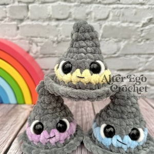 May include: Three gray crocheted wizard hats with different colored accents. The top hat has a yellow brim, the middle hat has a purple brim, and the bottom hat has a blue brim. Each hat has black eyes and a smiling mouth.
