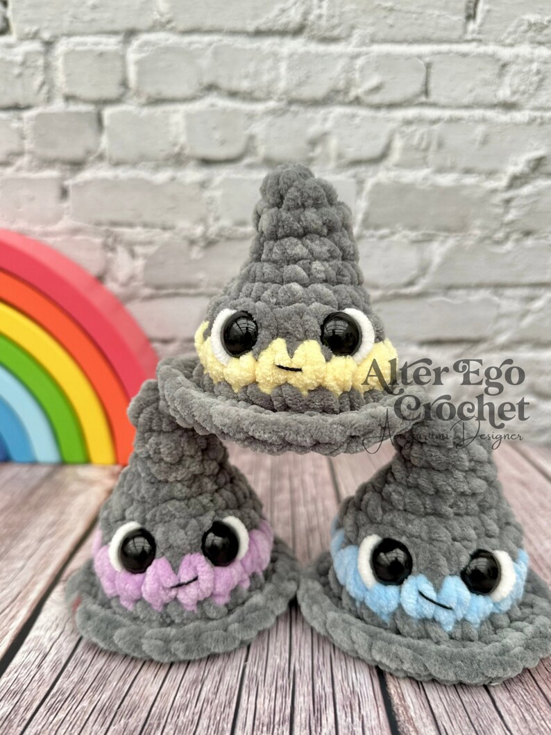 May include: Three gray crocheted wizard hats with different colored accents. The top hat has a yellow brim, the middle hat has a purple brim, and the bottom hat has a blue brim. Each hat has black eyes and a smiling mouth.