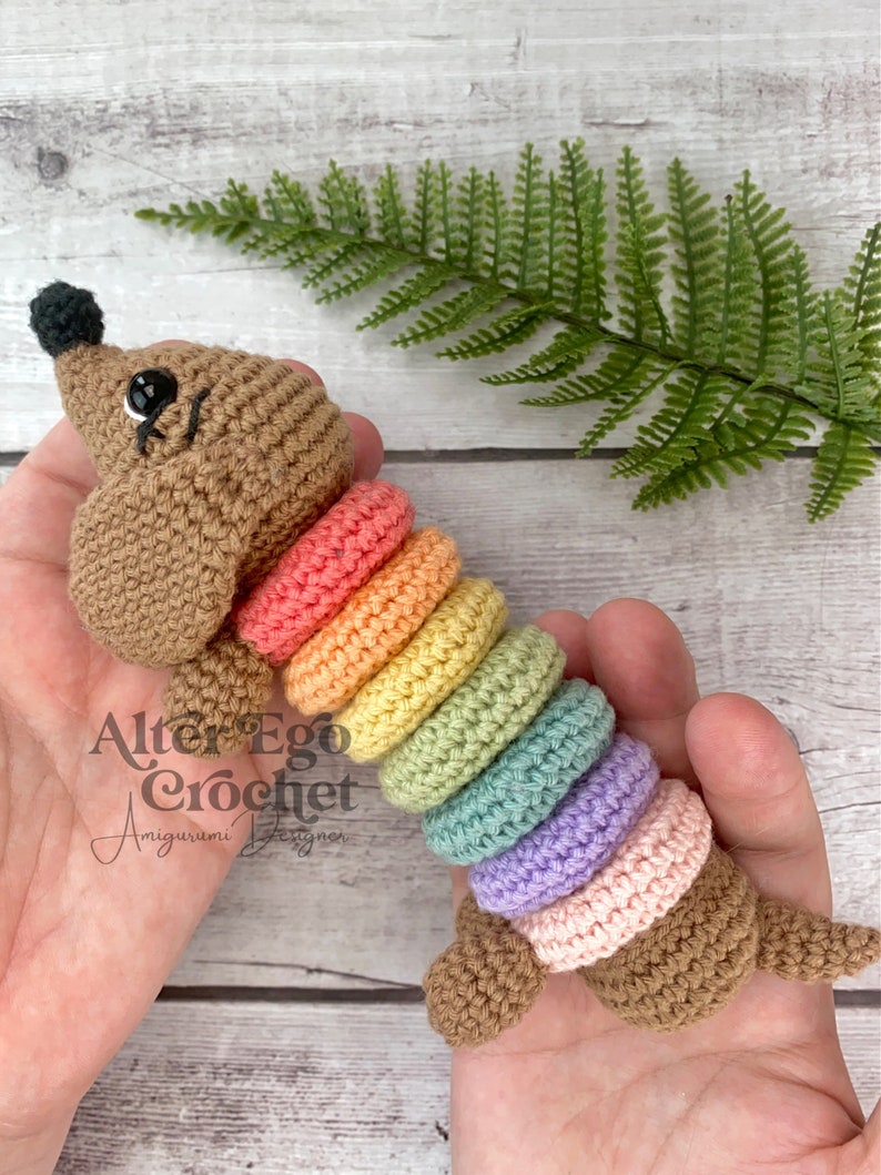 May include: A crocheted rainbow stackable toy with a brown crocheted dog head on each end. The toy is held in a person's hands. The text "Alter Ego Crochet Amigurumi Designer" is visible on the toy.