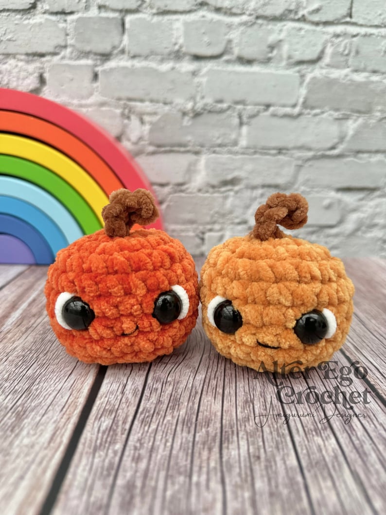 May include: Two crocheted pumpkins with smiling faces and googly eyes. The pumpkins are orange and brown and are sitting on a wooden surface.